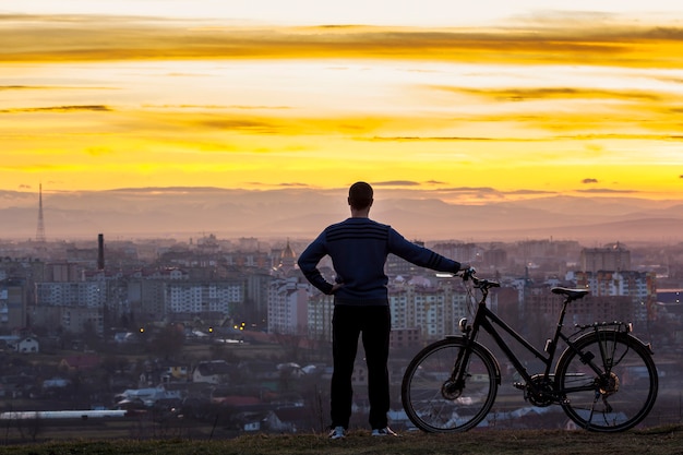 Dunkles Schattenbild eines Mannes, der nahe einem Fahrrad mit der Nachtstadtansicht steht