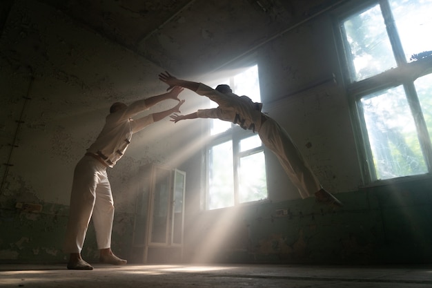 Dúo masculino haciendo trucos acrobáticos vistiendo trajes de locos en una habitación abandonada con rayos de sol desde las ventanas.