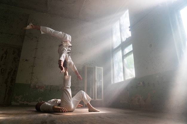 Dupla masculina fazendo truques acrobáticos vestindo fantasia de loucos em uma sala abandonada com os raios de sol das janelas.