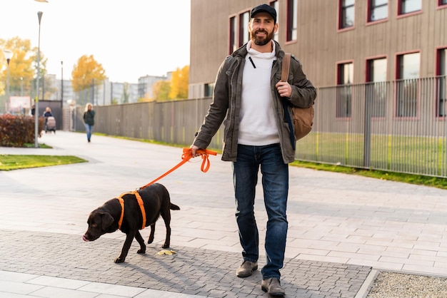 Durante a caminhada Proprietário de cachorro macho barbudo segurando uma coleira de seu cachorro Labrador enquanto passa um tempo juntos na rua Homem andando com seu animal de estimação Foto Premium