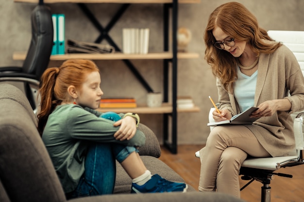 Durante a consulta profissional. Uma psicóloga simpática e simpática sentada perto de seu paciente enquanto faz anotações no caderno