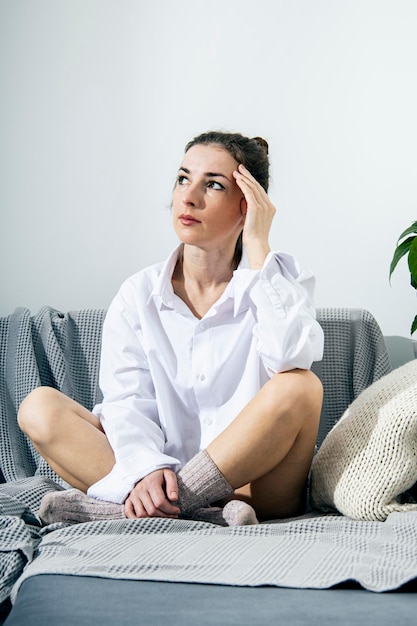 Durchdachte junge Frau im weißen Hemd und in den Socken, die auf Sofa sitzen