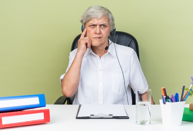 Durchdachte kaukasische Callcenter-Betreiberin auf Kopfhörern, die am Schreibtisch mit Bürowerkzeugen sitzen, die den Finger auf ihren Tempel legen und die Seite einzeln auf der grünen Wand betrachten