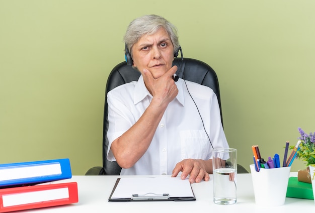 Durchdachte kaukasische Callcenter-Betreiberin auf Kopfhörern, die am Schreibtisch mit Bürowerkzeugen sitzen, die ihr Kinn halten