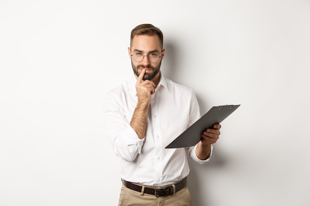 Durchdachter Büroangestellter, der Dokumente in der Zwischenablage hält und denkt, stehend über weißem Hintergrund.