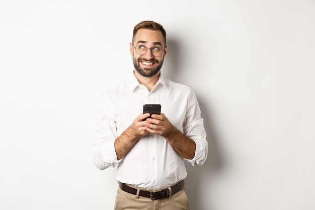 Durchdachter gutaussehender Manager, der Handy benutzt und darüber nachdenkt, eine Nachricht zu beantworten, in die obere linke Ecke schaut und lächelt, auf weißem Hintergrund stehend