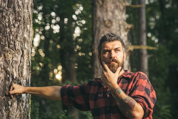 Durchdachter Mann, der nahe bei Baum steht. Neugieriger Holzfäller, der sein Kinn hält Hipster-Wanderung in der Natur