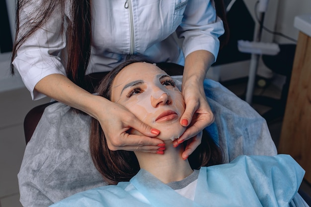 Durchführung von kosmetischen Dienstleistungen in einem Schönheitssalon. Eine junge attraktive Frau liegt auf einem Stuhl mit einer Stoffmaske mit nützlichen Substanzen.