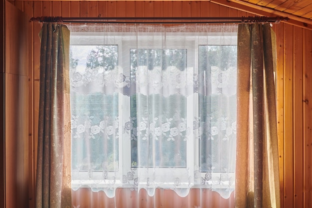 Durchscheinender weißer Vorhang im alten Stil am Fenster im Zimmer der Hütte. Fenster ist mit einem transparenten Vorhang bedeckt. Sonniges Morgenlicht im Hauszimmer.