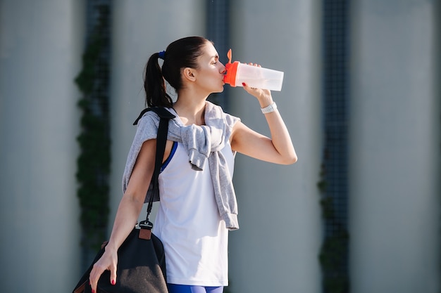 Durstige Frau im sportlichen Outfit steht auf der Straße und trinkt Wasser