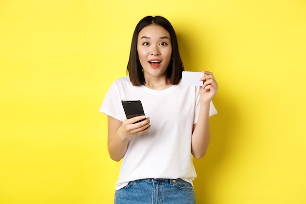 E-Commerce- und Online-Shopping-Konzept. Aufgeregte asiatische Frauenbestellung im Internet, Smartphone und Plastikkreditkarte haltend, über gelbem Hintergrund stehend.