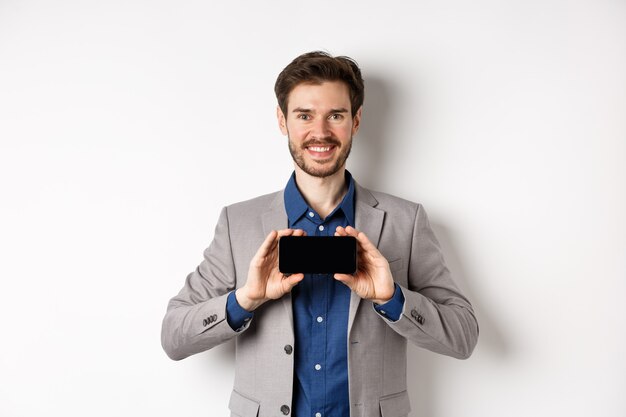 E-Commerce- und Online-Shopping-Konzept. Glücklicher Kerl im Geschäftsanzug, der leeren leeren Smartphonebildschirm horizontal zeigt, lächelnd erfreut an Kamera, weißer Hintergrund.