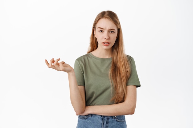 E daí. Mulher jovem confusa levanta uma mão e parece sem noção e questionada para a câmera, esperando por uma explicação, não consigo entender algo estranho, de pé sobre um fundo branco