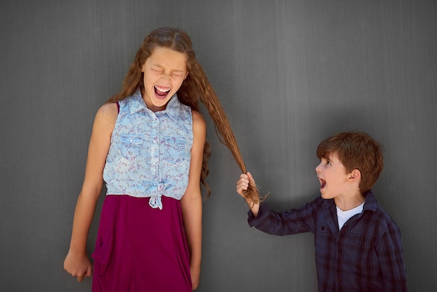 Foto É exatamente o que os irmãos mais novos fazem foto de estúdio de um menino puxando o cabelo da irmã enquanto está em pé contra um fundo cinza