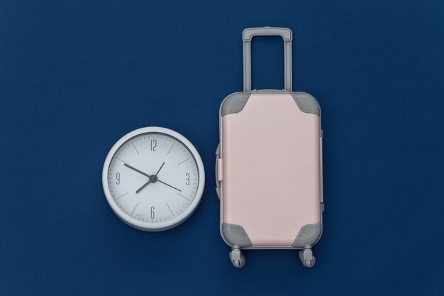 É hora de viajar. mini mala de viagem de plástico e relógio no fundo azul clássico. vista do topo. postura plana