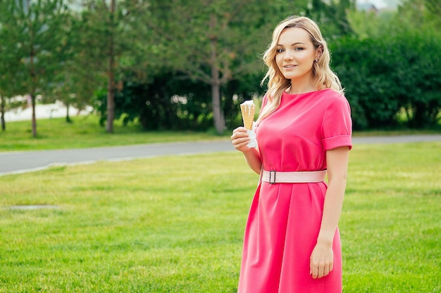 E linda jovem loira em um vestido rosa comendo sorvete de baunilha em um cone de waffle no parque