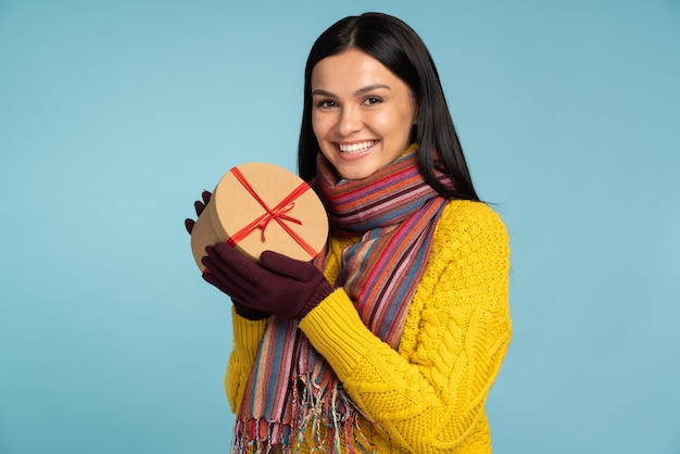 É para você. mulher linda sorridente no elegante suéter amarelo segurando a caixa de presente do armário em pé na frente da câmera e posando no fundo da parede azul. conceito de férias de inverno