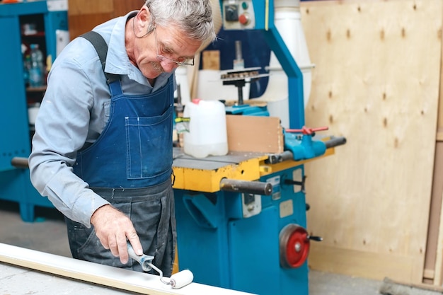 Un ebanista anciano con monos y gafas pinta una tabla de madera con un rodillo en un banco de trabajo en un taller de carpintería