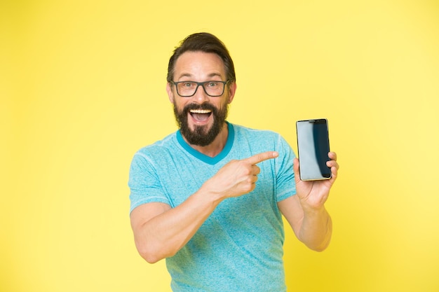 Echa un vistazo a la nueva aplicación. Guy anteojos alegres apuntando al teléfono inteligente. El usuario feliz del hombre recomienda probar la aplicación para teléfono inteligente. El hombre se beneficia de la comunicación online. Guy usuario de smartphone barbudo.