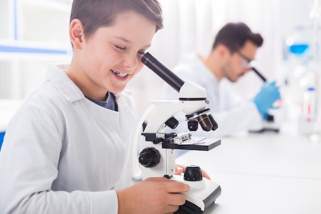 Echar un vistazo. Bonito niño inteligente tocando el microscopio mientras mira y sonríe