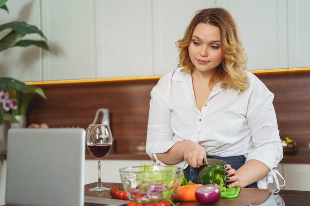 Echar un vistazo. Linda chica mirando la computadora, cocinando una cena saludable