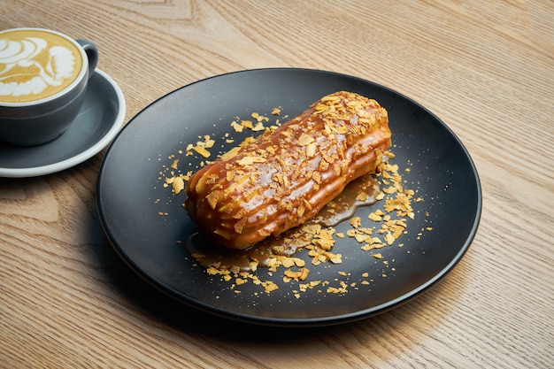 Eclair apetitoso y grande con glaseado de caramelo salado en la parte superior y crema de abeto en el interior en un plato negro. Dulces y postres para café y té.