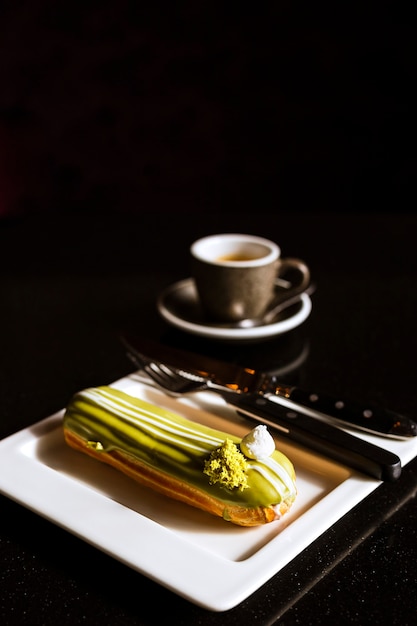 Eclair mit Pistaziencreme im Inneren auf einem Teller halbiert und eine Tasse Kaffee auf dunklem Tisch.