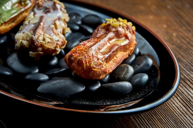 Eclairs franceses con natillas y coberturas variadas servidas en una placa negra sobre un fondo de madera. Comida de restaurante.