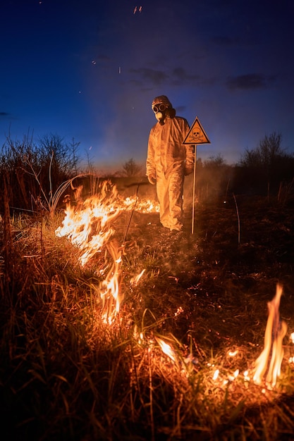 Ecologista apagando incêndio em um campo à noite Homem de terno e máscara de gás perto de grama em chamas com fumaça e triângulo amarelo com crânio e ossos cruzados sinal de alerta Conceito de desastre natural