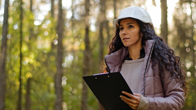 Ecologista técnica milenaria mirando hacia las copas de los árboles Joven india en casco con portapapeles tomando medidas revisando árboles Ingeniero forestal en el parque Supervisando el santuario de vida silvestre