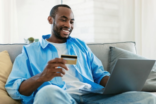 Ecommerce Feliz homem negro segurando cartão de crédito e compras on-line com laptop sentado no sofá em casa