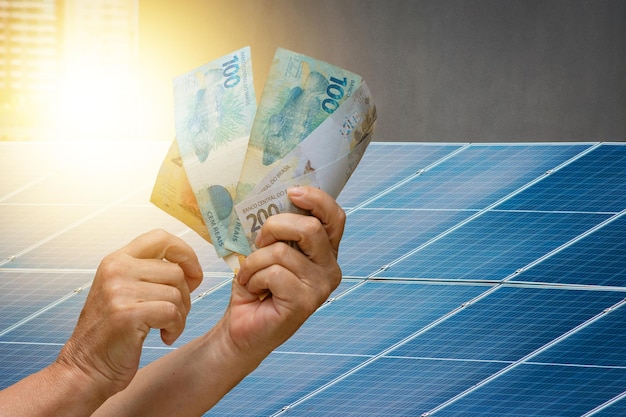 Economía de energía solar, sosteniendo dinero brasileño frente al panel fotovoltaico en un techo, fondo de puesta de sol. Espacio para texto.