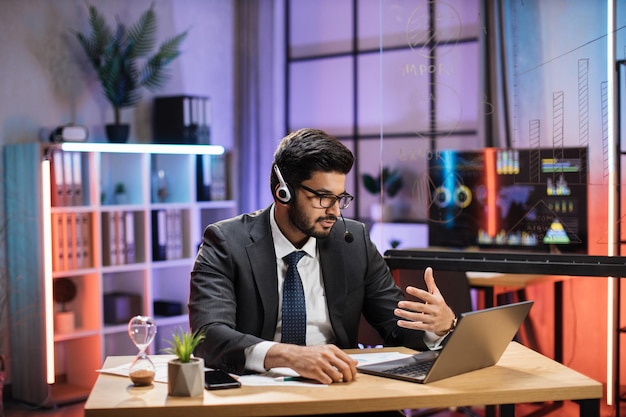 Economista empresário indiano experiente em terno usando fone de ouvido sentado à mesa com laptop