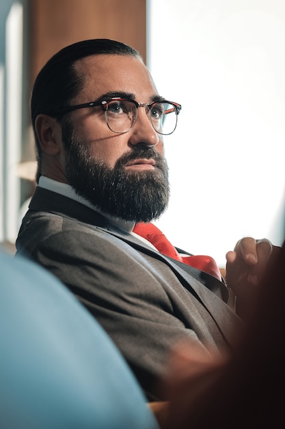 Economista con gafas. Economista barbudo de pelo oscuro con gafas sentado en el sofá de su habitación de hotel