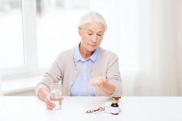 edad, medicina, atención médica y concepto de personas - mujer mayor con pastillas y vaso de agua en casa