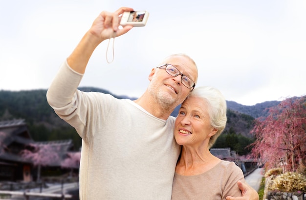 edad, turismo, viajes, tecnología y concepto de personas - pareja mayor con cámara tomando selfie sobre el fondo del paisaje de la aldea asiática