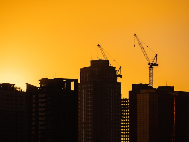 Edificio bajo grúas de construcción y silueta de edificio de gran altura en el atardecer