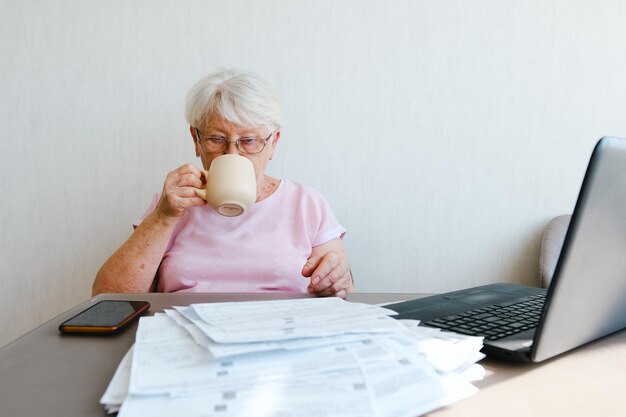 Editor maduro leyendo texto impreso haciendo notas usando una computadora portátil trabajando desde casa tomando café té