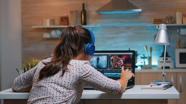 Editor de video con auriculares y trabajando desde casa en un proyecto digital sentado en la cocina. Videógrafo de montaje de película de audio de edición en portátil profesional sentado en el escritorio a medianoche