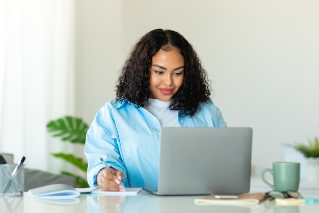 Educação a distância mulher negra obesa sentada na mesa usando laptop e escrevendo no caderno estudando online em casa