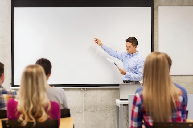 educação, ensino médio, tecnologia e conceito de pessoas - professor com bloco de notas, computador portátil em frente aos alunos e mostrando algo no quadro branco em sala de aula