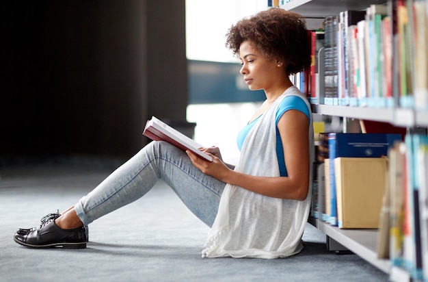 educação, ensino médio, universidade, aprendizado e conceito de pessoas - estudante afro-americana lendo livro sentado no chão da biblioteca