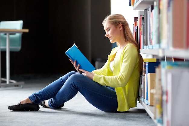 educação, ensino médio, universidade, aprendizado e conceito de pessoas - estudante sorridente lendo livro sentado no chão da biblioteca
