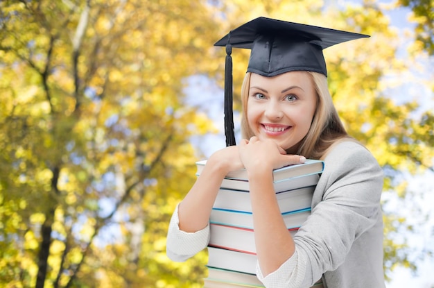 Educação, escola, conhecimento, graduação e conceito de pessoas - estudante sorridente feliz em chapéu de solteiro com livros sobre fundo de árvores de outono