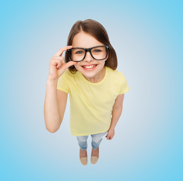 Foto educação, escola, pessoas, infância e conceito de visão - menina sorridente em óculos pretos sobre fundo azul