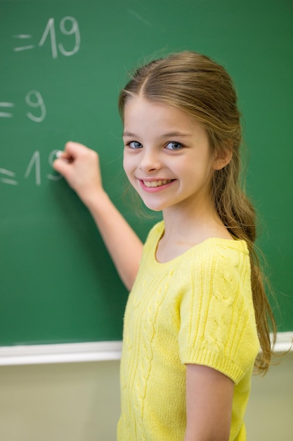 Educação, escola primária, aprendizagem, matemática e conceito de pessoas - garotinha sorridente escrevendo números no quadro de giz verde em sala de aula