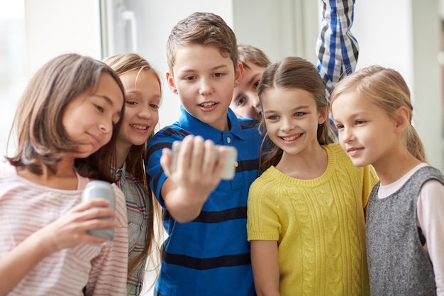 educação, escola primária, bebidas, crianças e conceito de pessoas - grupo de crianças da escola com smartphone e latas de refrigerante tomando selfie no corredor