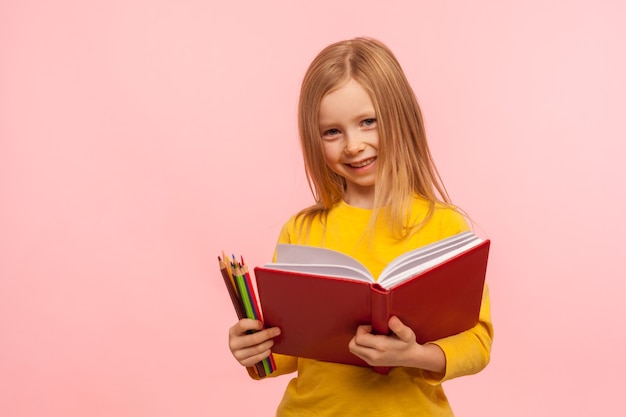 Educação, escola primária. retrato de uma menina adorável sorrindo para a câmera, segurando um grande livro e lápis, desenvolvimento de habilidades criativas da criança. foto de estúdio interno isolada em fundo rosa