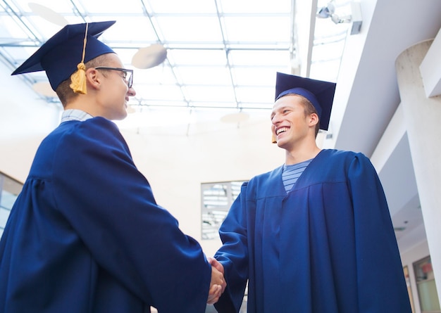 educação, graduação e conceito de pessoas - estudantes sorridentes em capelos e vestidos apertando as mãos ao ar livre