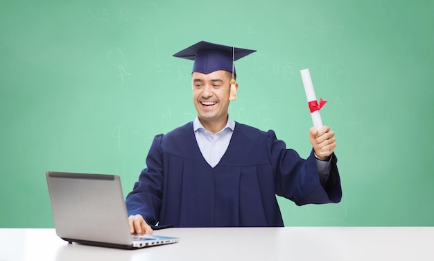 educação, graduação, tecnologia e conceito de pessoas - estudante adulto feliz em capelo com diploma e computador portátil sentado à mesa sobre fundo de quadro de giz escola verde